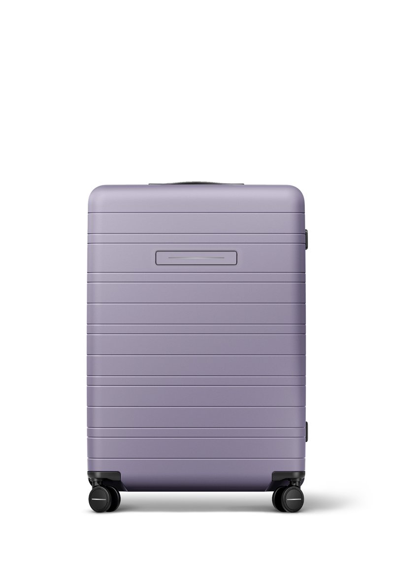 Valise rigide de couleur violet clair, avec une texture nervurée et une poignée supérieure, équipée de quatre roues noires pour une mobilité facile.