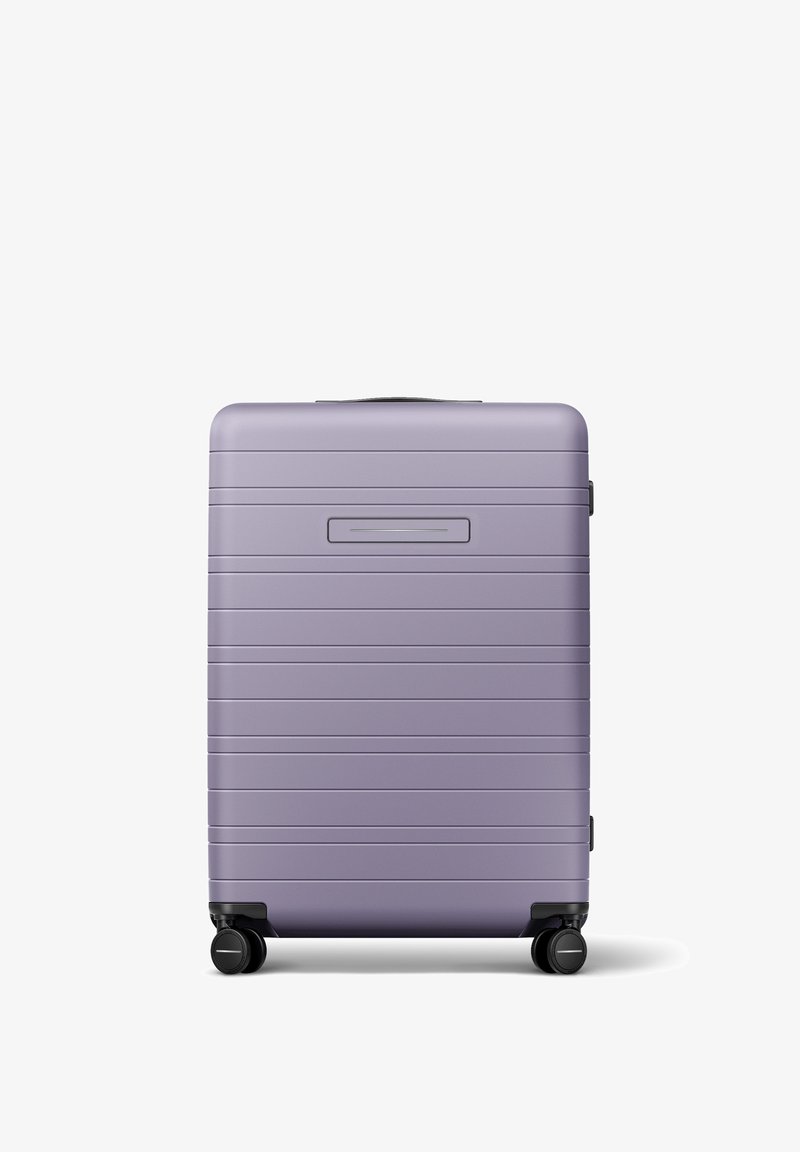 Valise rigide de couleur violet clair, avec une texture nervurée et une poignée supérieure, équipée de quatre roues noires pour une mobilité facile.