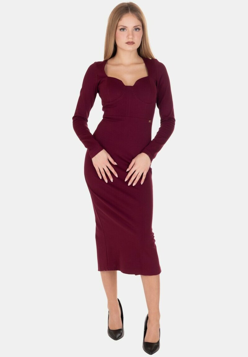 Vestito aderente color borgogna a maniche lunghe con scollo quadrato e orlo fino alla coscia, realizzato in un tessuto liscio ed elastico. Completa il look un paio di tacchi neri.
