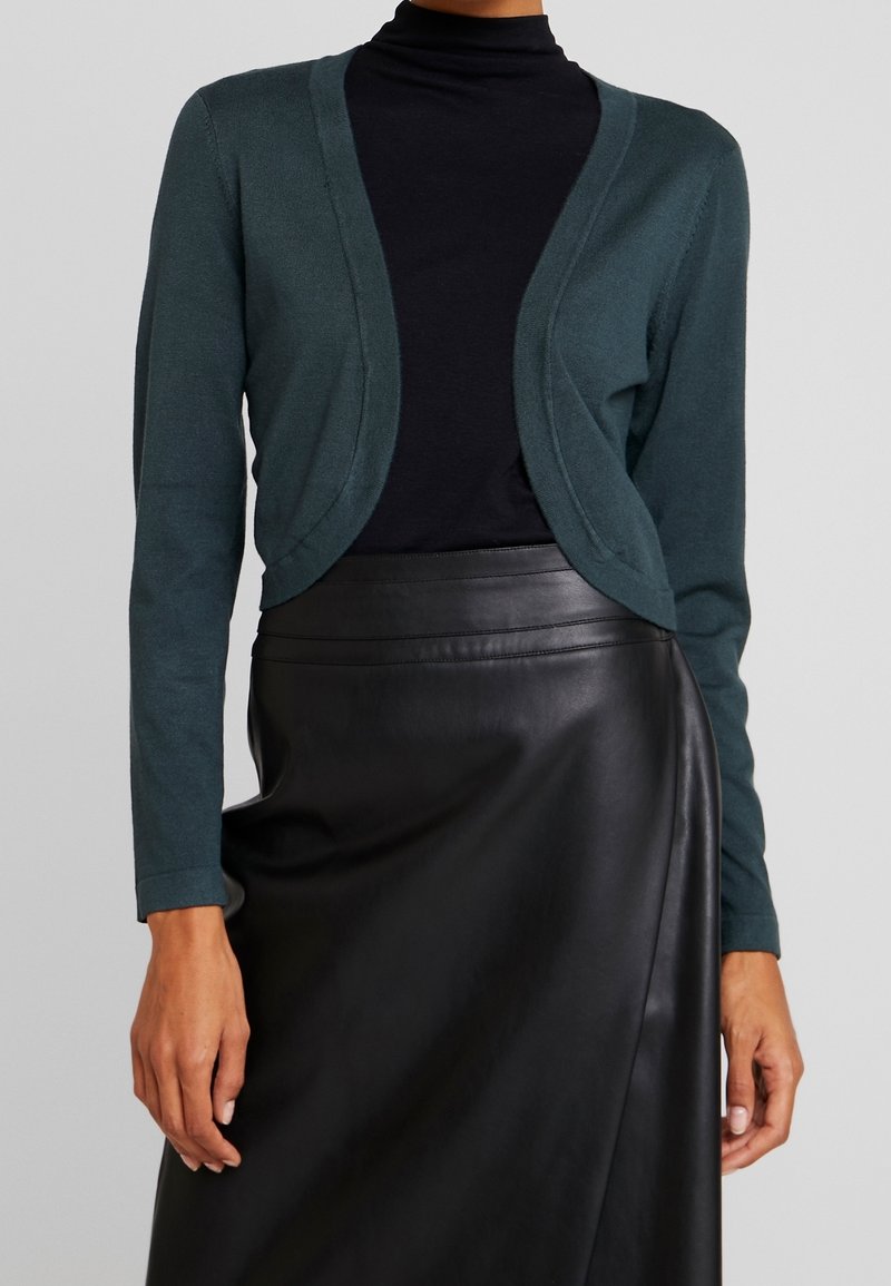 Woman wearing dark green cropped cardigan over black turtleneck and black high-waisted leather skirt against plain background.