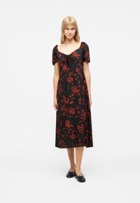 Jeune femme debout portant une robe midi noire à imprimé floral rouge, manches bouffantes, encolure carrée, et des ballerines noires sur fond blanc.
