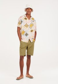Camisa de manga corta con un patrón geométrico multicolor, combinada con pantalones cortos oliva y chanclas marrones. Accesorios incluyen un sombrero de cubo blanco.