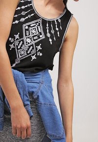 Haut noir sans manches avec des motifs géométriques blancs brodés et des accents. Associé à une veste en denim bleu clair nouée à la taille.