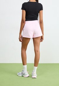 Černé přiléhavé tričko, růžové sportovní kraťasy s elastickým pasem a šedobílé tenisky s růžovými akcenty. Pozadí je zelené.