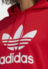 Röd hoodie med en stor vit adidas-logga som har tre bladliknande element och tre horisontella ränder. Tillverkad av bomull med dragsko.