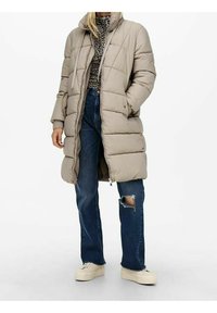 Kvinde iført en beige lang pufferjakke, leopardmønstret top, slidte blå jeans og hvide sneakers, stående mod en hvid baggrund.