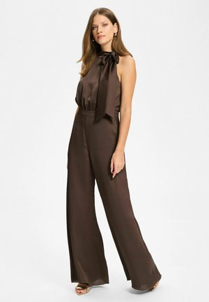 Femme portant une combinaison sans manches en satin brun foncé avec col haut et nœud latéral, associée à des sandales dorées et des boucles d'oreilles créoles.