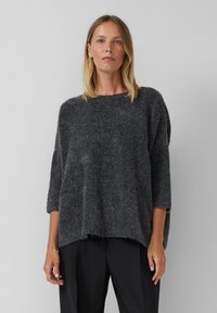 Femme portant un pull moelleux gris foncé ample avec des manches trois-quarts et un pantalon noir, se tenant devant un arrière-plan gris clair uni.