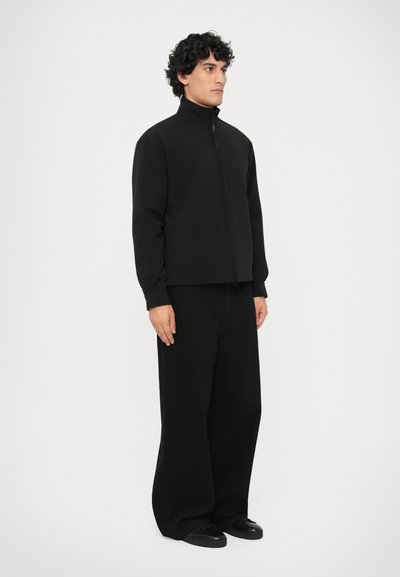 Čierna bunda na zips s vysokým golierom, skombinovaná s voľnými čiernymi nohavicami. Tkanina vyzerá hladko. Model stojí na bielom pozadí.