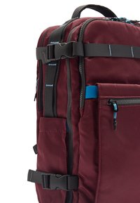Bordowy plecak z czarnymi paskami i elementami metalowymi, zapinaną na zamek kieszenią z niebieskimi akcentami oraz fakturowaną powierzchnią materiału. Widać regulowane klamry.
