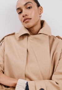 Beige trenchcoat met een hoge kraag en schouderepauletten, met gemerkte mouwen met een gebreide accent. Gladde katoenstof.