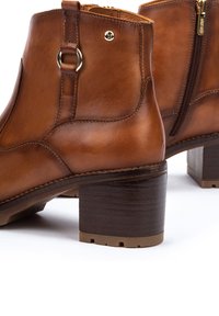 Botas de tobillo de cuero marrón con puntera redonda, cremallera lateral, herrajes metálicos decorativos y un tacón de bloque en un tono marrón oscuro contrastante.