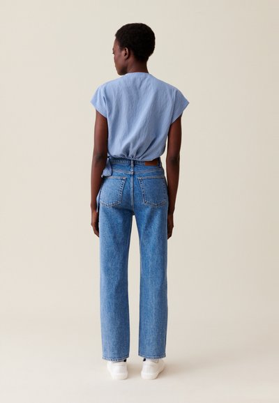 Femme de dos portant un blouse légère à manches courtes bleu clair, un jean droit taille haute bleu et des baskets blanches sur un fond beige.