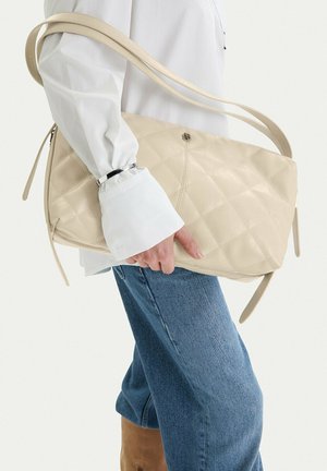 Osoba v modrých džínách a bílém tričku držící velkou béžovou prošívanou koženou kabelku s dvojitými popruhy.