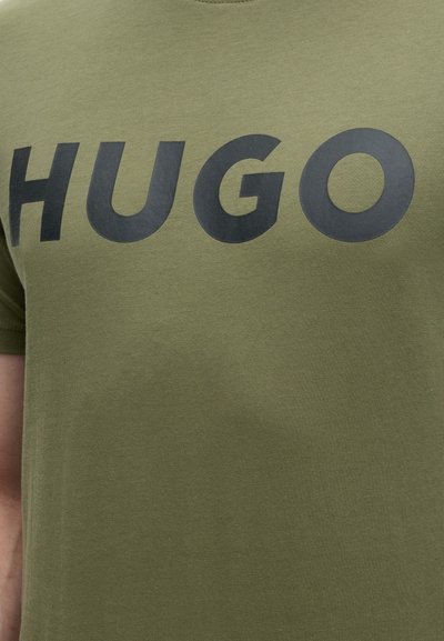 Тъмнозелена риза с голям черен надпис "HUGO" отпред на гърдите, носена от човек.