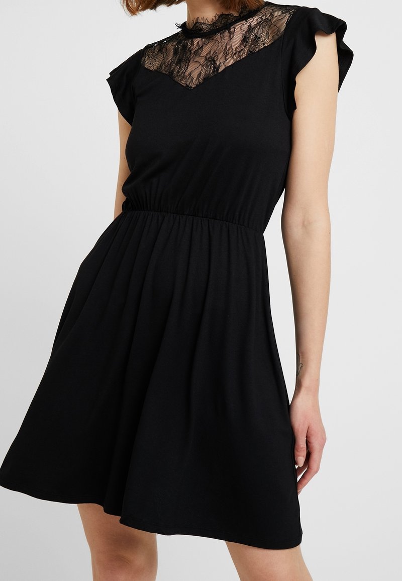 Robe noire sans manches avec empiècement en dentelle, col montant, volants aux épaules et taille froncée portée par une personne debout.
