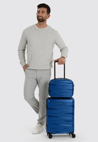 Man in een lichtgrijs outfit staat met hand op de handgreep van bijpassende blauwe harde koffers, een grote en een kleine, op een witte achtergrond.