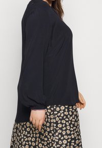 Langsleeve, marineblauwe blouse met een losse pasvorm, voorzien van een ronde halslijn en subtiele, geprinte afwerking. Gekleed over een zwarte rok met bloemenpatroon.