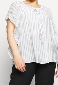 Túnica blanca con rayas verticales grises, escote redondeado y detalle de cordón trenzado en el escote. Tela suelta y fluida.