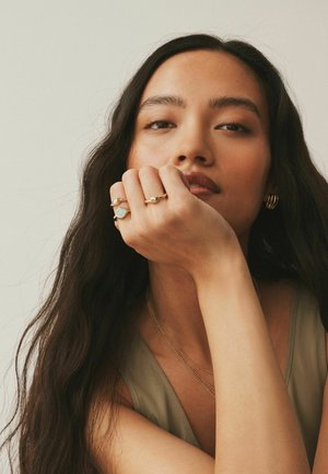 Gold-tone rings with turquoise and textured accents worn on a hand resting against the face. Long, dark hair frames the face.