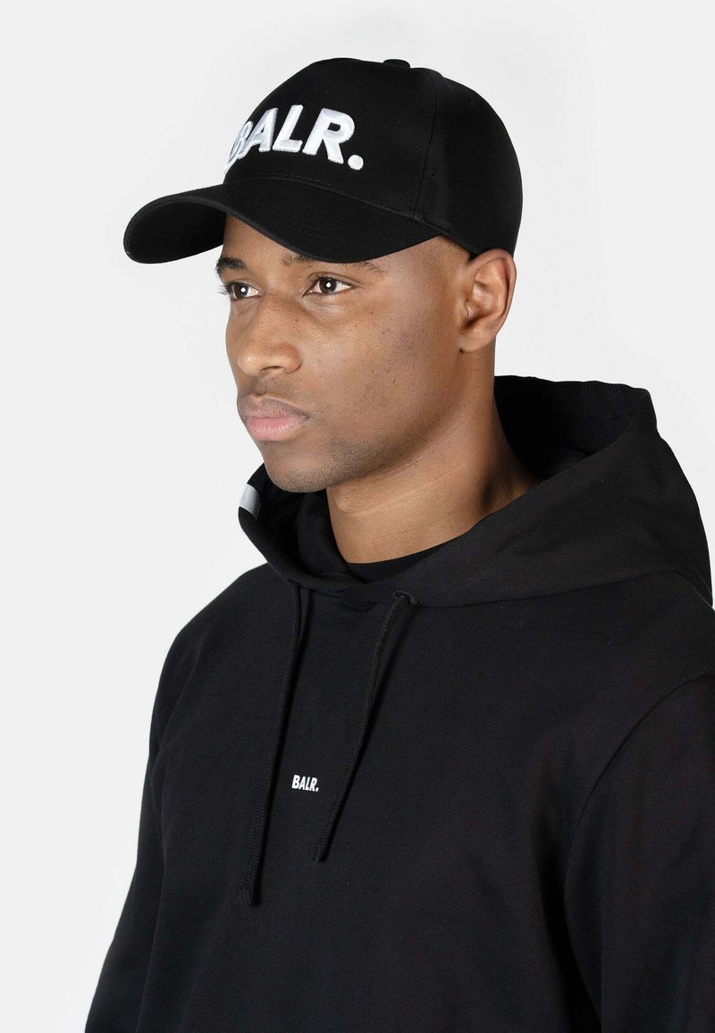 Cască neagră cu logo alb "BALR." pe față, având o vizieră curbată. Purtați-o cu un hanorac negru. Textura netedă a țesăturii, design minim.