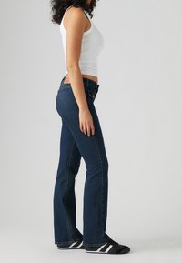 Jean évasé bleu foncé en denim, avec une taille mi-haute, cinq poches et un motif de couture classique. Porté avec un débardeur blanc et des chaussures noires.