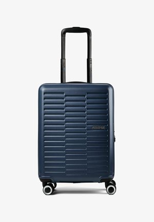 Maleta rígida azul marino con relieves horizontales, asa negra retráctil, cuatro ruedas giratorias y logo de American Tourister en el frontal.