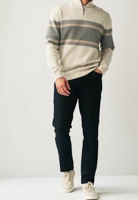 Maglione beige con zip e collo a lupetto, caratterizzato da righe orizzontali grigie e pesca, abbinato a pantaloni neri e sneakers di colore chiaro. Tessuto uniforme e morbido.