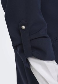 Chemise bleu marine avec manches retroussées et fermeture par bouton, doublure intérieure blanche. Tissu lisse avec une coupe ajustée et des poignets nets.