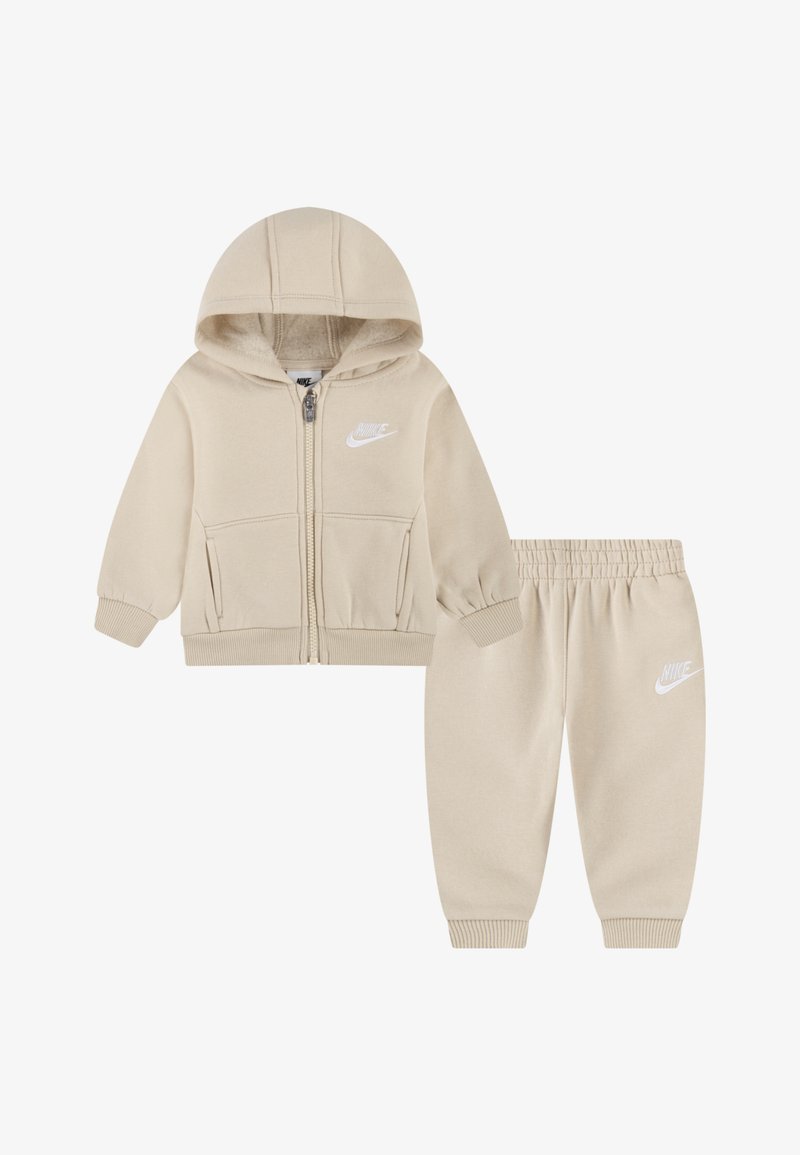 Beige zip-up hoodie med hætte og forlommer, parret med matchende bukser med elastisk talje. Begge har et hvidt Nike-logo. Blødt stof.