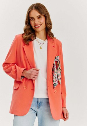 Korálový blazer so strihom na mieru, na dva gombíky a s kvetinovou vreckovou šatkou. Pod ním je biela košeľa, doplnená svetlomodrými džínsy.