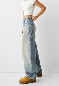 Vaqueros de pierna ancha en azul claro con un acabado desgastado y una cintura alta, combinados con botas amarillas de suela gruesa. Sin patrones ni acentos visibles.