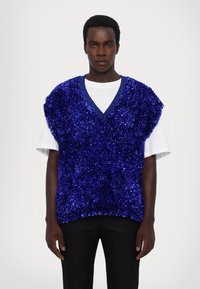 Mėlynas blizgučių liemenė su V formos iškirpte, uždėta ant baltos marškinėlių. Trumpi rankoviai, plunksnuotas tekstūras ir laisvas kirpimas. Nešiojama su juodomis kelnėmis.