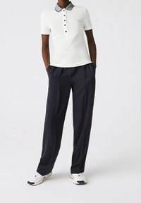 Polo shirt blanc avec un col foncé, des boutons noirs, associé à un pantalon plissé bleu marine. Des baskets blanches avec des accents rouges complètent la tenue.