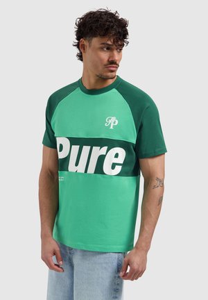 Mann trägt grün-tealfarbenes T-Shirt mit dem Schriftzug „Pure“, hellblaue Jeans und sichtbare Armtattoos, schaut zur Seite.