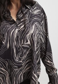 Blusa a maniche lunghe in tessuto nero con un motivo a onde beige fluttuante. Presenta bottoni lungo il davanti e un colletto classico.