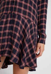 Marinblå rutig klänning med röda och vita rutmönster, knäppning framtill, långa ärmar och en utsvängd nederkant i en texturerad tyg.