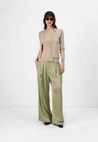 Beige Strickjacke mit Knöpfen, über einem passenden Oberteil getragen, kombiniert mit lockeren olivgrünen Hosen. Das Model trägt außerdem rote Sonnenbrillen.