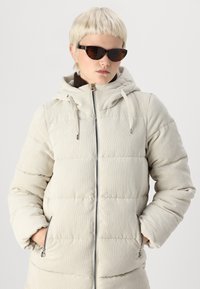 Doudoune en velours côtelé beige clair, dotée d'une capuche, d'une fermeture éclair et de coutures verticales. Agrémentée de poches avant et de cordons de serrage.