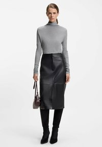 Vrouw in een strak grijze coltrui, zwarte leren midi rok, zwarte kniehoge laarzen en met een kleine donkere handtas.