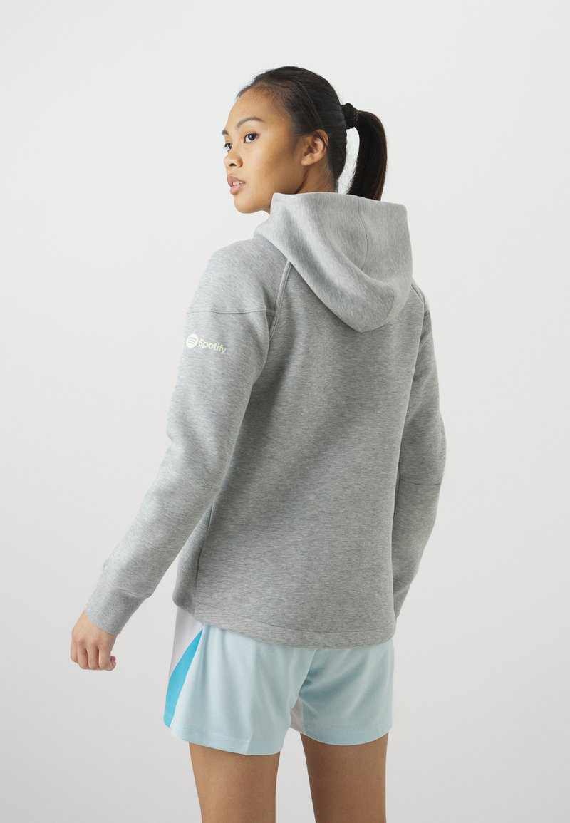 Szara bluza z kapturem i kieszenią z przodu, z małym zielonym logo Spotify na rękawie. Noszona z jasnoniebieskimi szortami sportowymi.