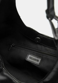 Zwarte leren handtassengebied met een gladde textuur, een ritssluitingvak en een label met de tekst "PULL&BEAR." Rond drukknopsluiting zichtbaar.