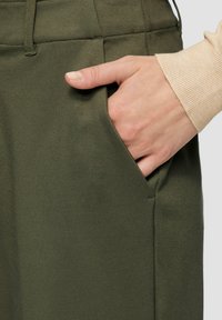 Olijfgroen stoffen broek met een relaxte pasvorm. Kenmerkt zich door een zijzak met een gladde textuur. Hand rustend in de zak, waarbij het ontwerp wordt getoond.