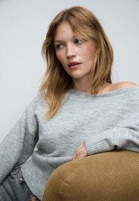 Pull en tricot gris à côtes avec un design décolleté, associé à un jean décontracté ; l'arrière-plan présente une atmosphère neutre et claire.