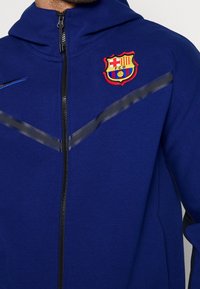 Felpa con zip blu e cappuccio, caratterizzata da una cerniera nera, strisce nere in rilievo e un logo ricamato del Barcelona FC sul petto.