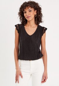 Haut noir à manches courtes avec un décolleté en v et un tissu texturé. Présente des bordures en dentelle sur les manches et une coupe décontractée, porté avec un pantalon blanc.
