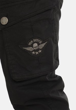 Poche sur un pantalon cargo noir avec un écusson représentant un crâne ailé, trois étoiles, et le texte « Von Dutch ».