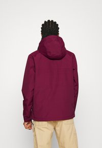 Veste imperméable bordeaux avec capuche, poignets élastiques et ourlet à cordon de serrage. Texture lisse, design simple, sans logo ni accent visible.