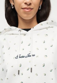Κοντινό πλάνο ενός ατόμου που φοράει λευκό hoodie με μικρά λουλουδάτα σχέδια και το κέντημα "Guess Jeans" σε καλλιγραφικά γράμματα στο στήθος.