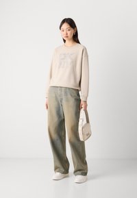 Bežinė sportinė maikutė su tekstūruotu "DKNY" logotipu, derinama su plačiomis išblukusiomis mėlynomis džinsomis. Modelis laiko šviesiai rudos spalvos petinį krepšį.